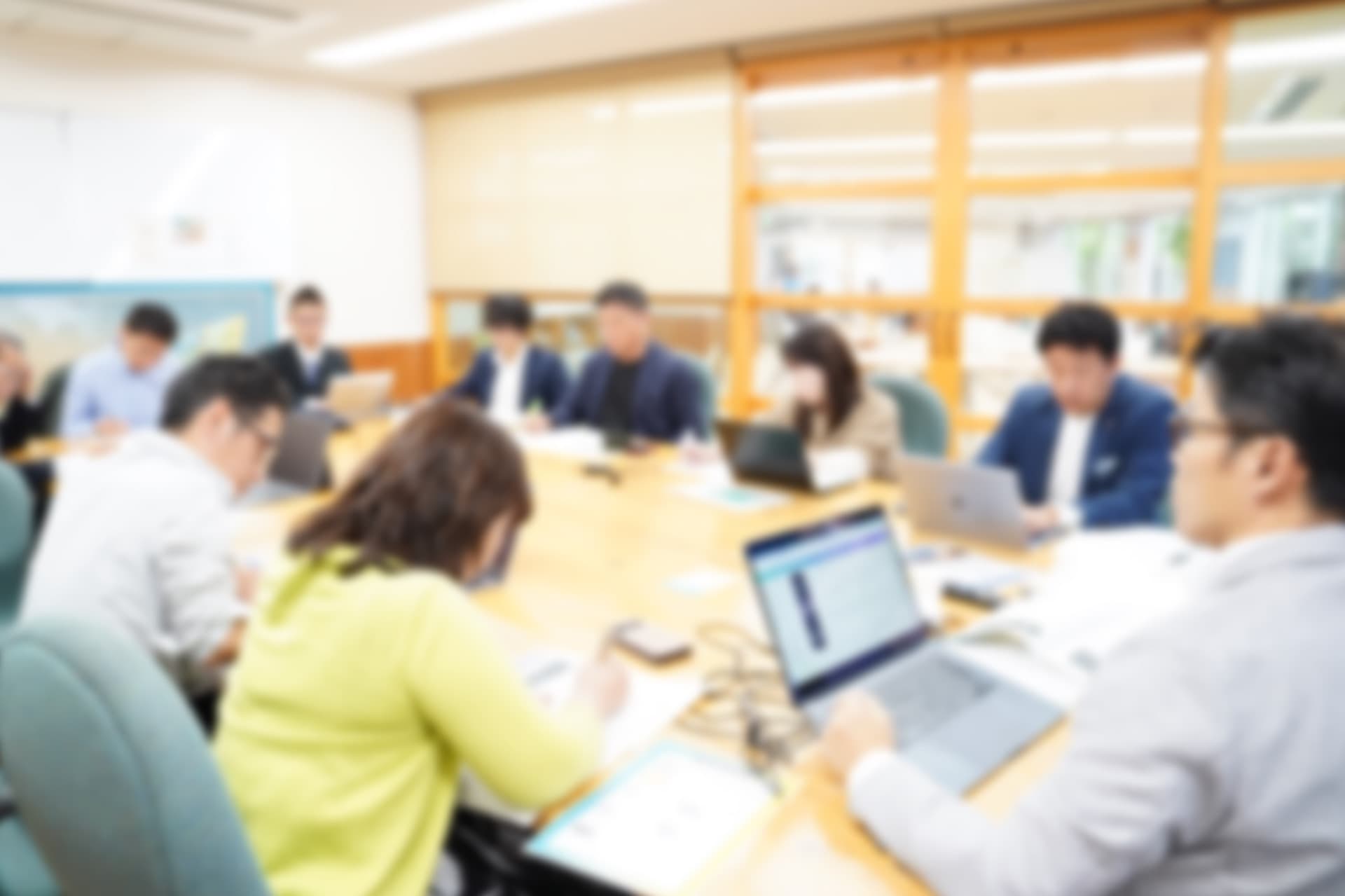 セミナー会場で、経営者たちが大きな机を囲みノートパソコンと資料を広げて学んでいる風景(ぼかし加工)